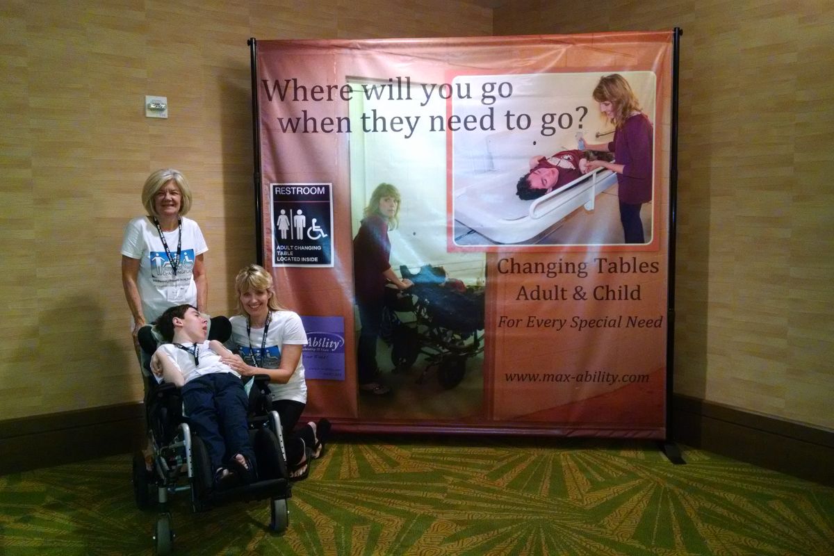 Sabrina Kimball, her son Greyson Channing, and Deretha Heller advocating for Universal Changing Places at The Family Café, standing beside a large promotional banner sponsored by MaxAbility.
